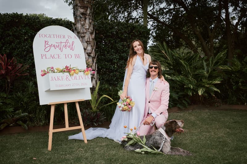 Bride Olivia and groom Jake pose for a couple portrait at Eatons Hill Hotel with their dog lying on the grass. Olivia is standing in a white wedding dress holding a bouquet, while Jake kneels beside her in a pink suit and sunglasses. A wedding sign with floral decoration reads 'Together is a Beautiful Place to Be, Jake & Olivia, February 27th, 2026'.
