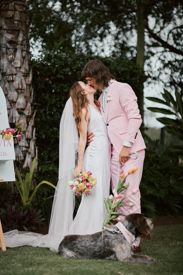 The bride Olivia and groom Jake share a kiss during their couple portraits at Eatons Hill Hotel, with Olivia holding a bouquet and a dog lying on the grass in front of them.