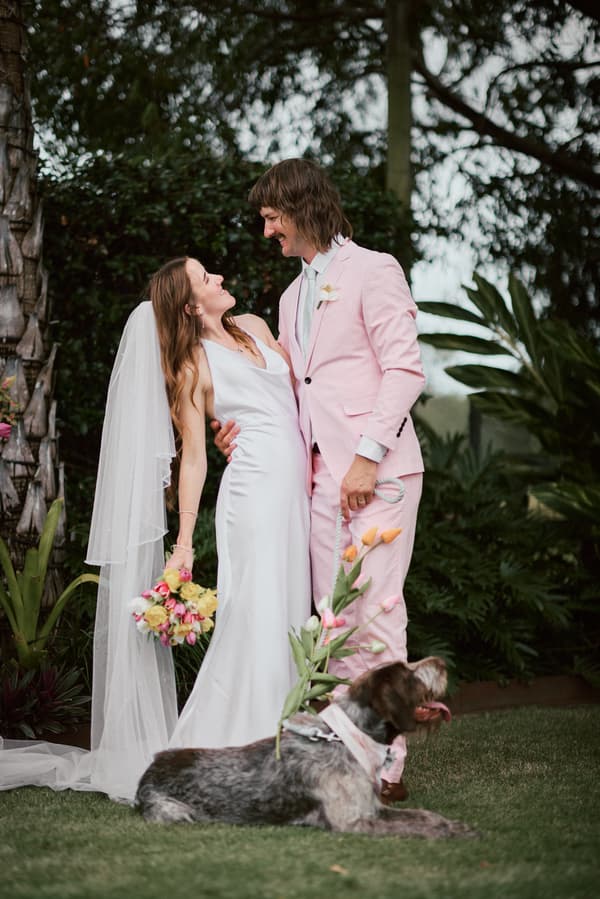 Olivia the bride in a white gown and veil holding a bouquet stands with Jake the groom in a light pink suit at Eatons Hill Hotel, with their dog lying on the grass in front of them holding flowers in its mouth.