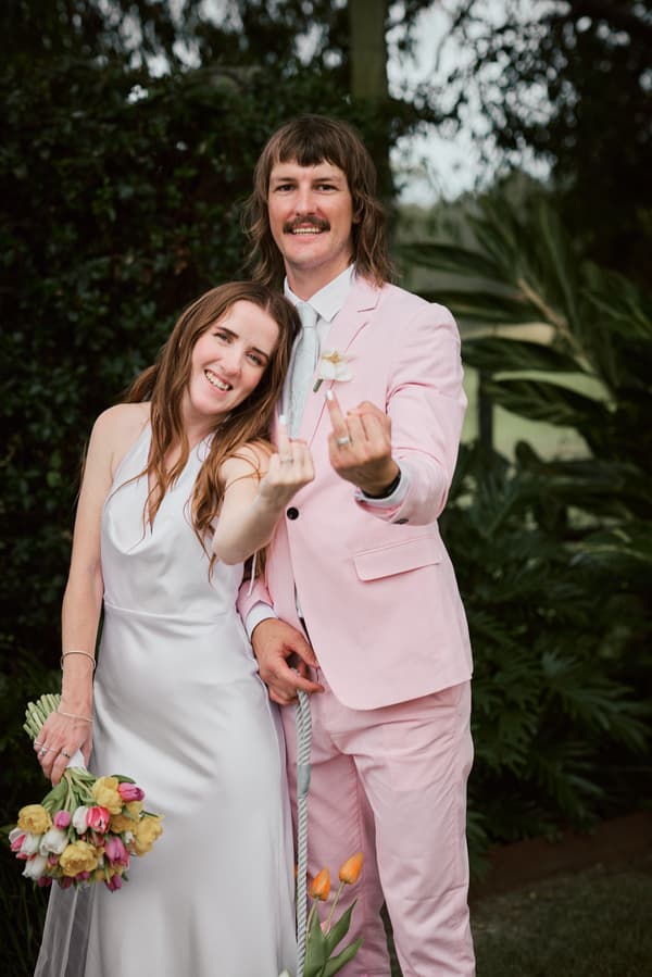 Bride Olivia in a white wedding dress holding a bouquet of tulips and groom Jake in a pink suit with a white tie pose together outdoors at Eatons Hill Hotel, both showing their wedding rings on their middle fingers.