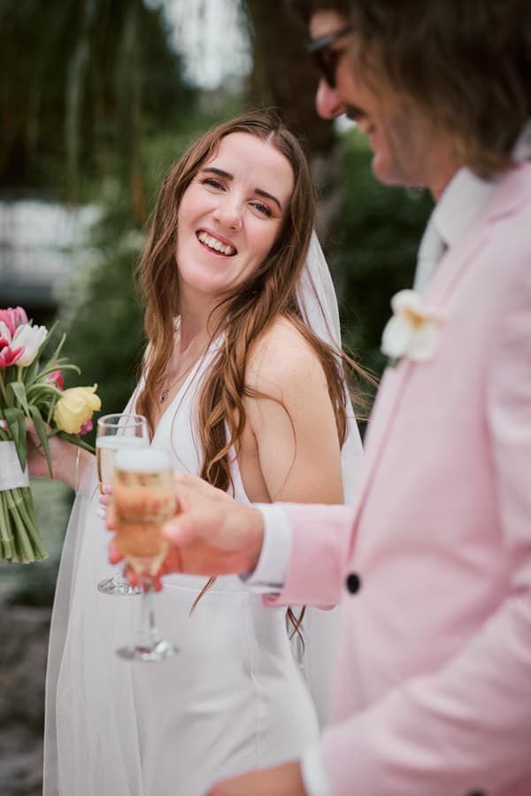 The bride Olivia in a white wedding dress holding a bouquet of tulips and a glass of champagne, smiling towards the groom Jake, who is wearing a light pink suit and holding a glass of champagne, at Eatons Hill Hotel.
