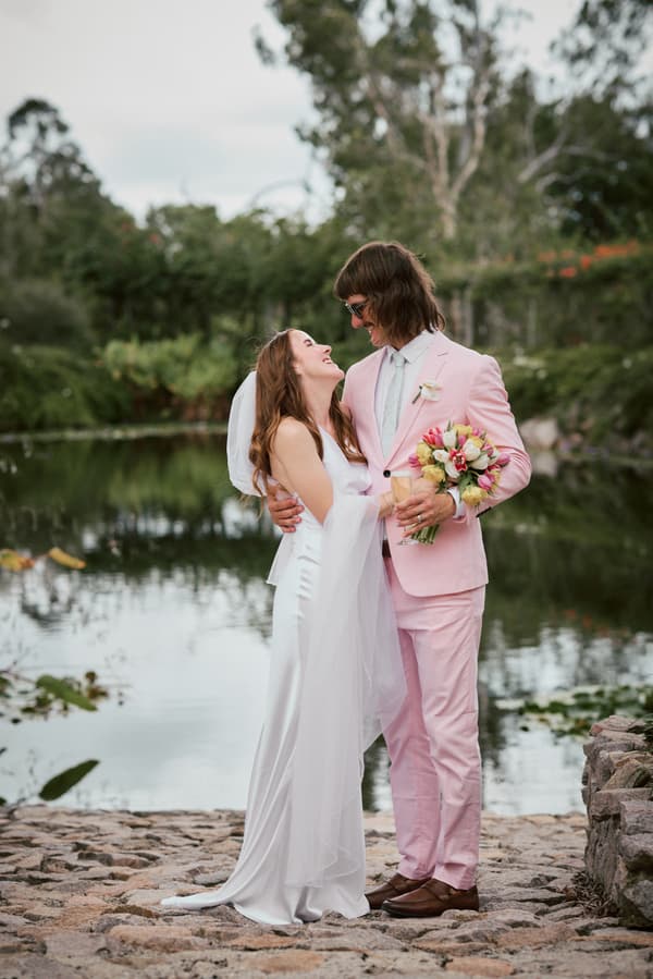 The bride Olivia in a white wedding gown and veil embraces the groom Jake, who is wearing a light pink suit and holding a bouquet of flowers, standing on a stone path by a pond at Eatons Hill Hotel.