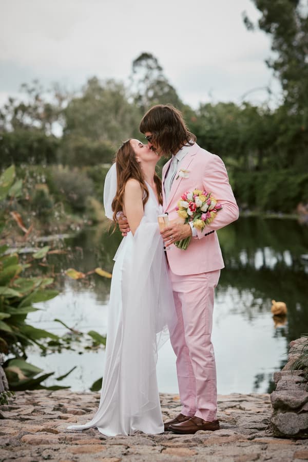 Olivia the bride and Jake the groom kiss while standing on a stone path beside a pond at Eatons Hill Hotel. Olivia wears a white wedding dress and veil, and Jake wears a light pink suit holding a bouquet of flowers and a glass.