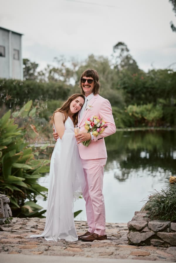 The bride Olivia in a white wedding dress and the groom Jake in a pink suit stand embracing beside a pond at Eatons Hill Hotel, with Jake holding a bouquet of flowers and a champagne flute.