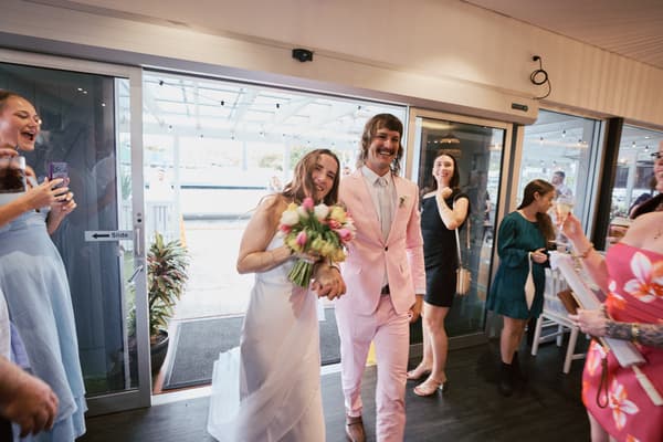 Bride Olivia holding a bouquet and groom Jake in a pink suit walk hand in hand inside White Horse Ranch reception area, surrounded by guests taking photos and holding drinks.