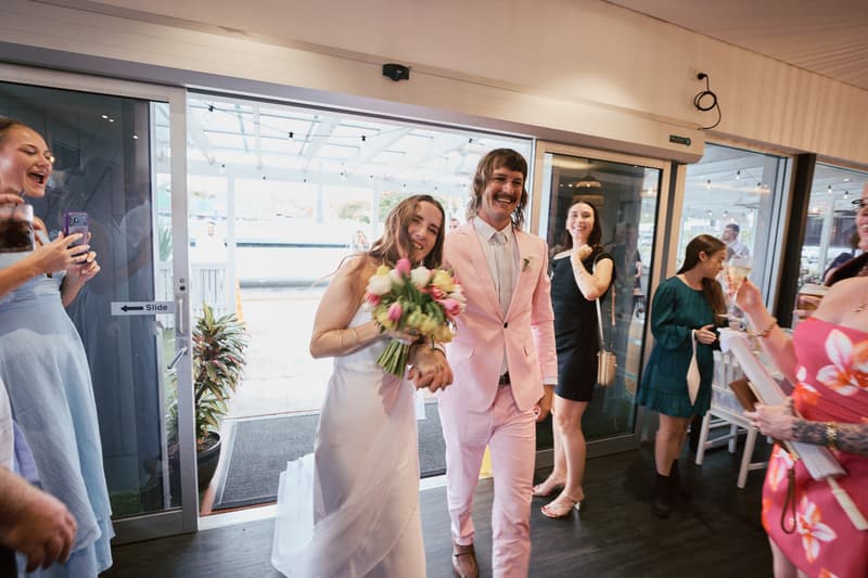Bride Olivia holding a bouquet and groom Jake in a pink suit walk hand in hand inside White Horse Ranch reception area, surrounded by guests taking photos and holding drinks.