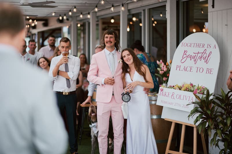 Jake and Olivia stand together at the reception stage at White Horse Ranch, with Olivia holding a clock and Jake in a pink suit. Guests are visible in the background, and a sign reads 'Together is a Beautiful Place to Be - Jake & Olivia - February 27th, 2026.'