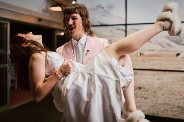Jake carries Olivia in his arms at the reception stage of White Horse Ranch, both dressed in wedding attire with Olivia wearing slippers.