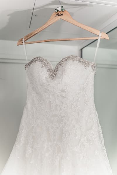 The bride's white lace wedding dress with beaded detailing is hanging on a wooden hanger labeled 'Bride'. Brisbane videographer by She Said Yes.