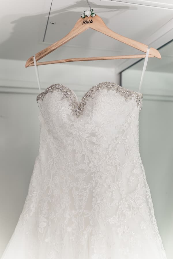 The bride's white lace wedding dress with beaded detailing is hanging on a wooden hanger labeled 'Bride'.