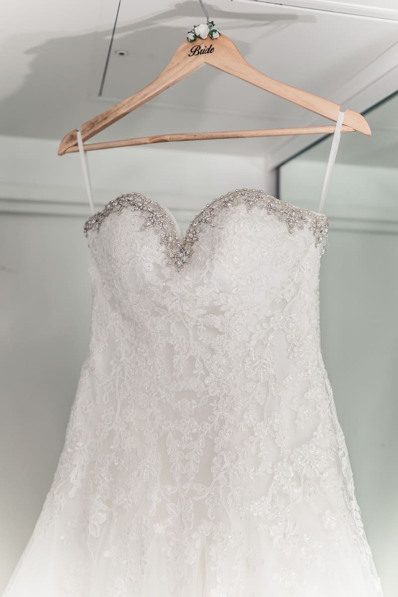 The bride's white lace wedding dress with beaded detailing is hanging on a wooden hanger labeled 'Bride'. Brisbane videographer by She Said Yes.