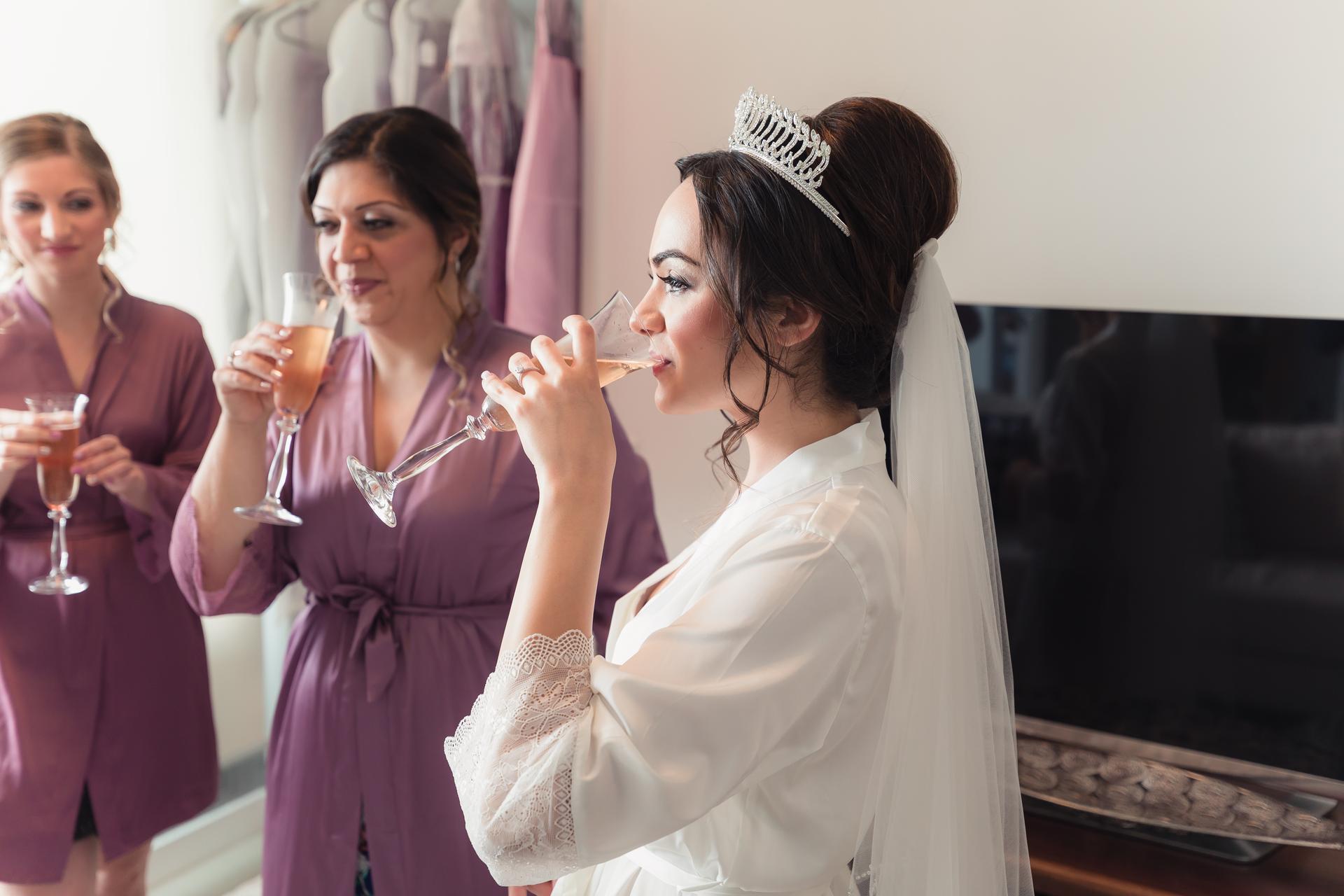The bride Maryam wearing a tiara and veil drinks from a glass while two bridesmaids in matching purple robes hold glasses in the background.