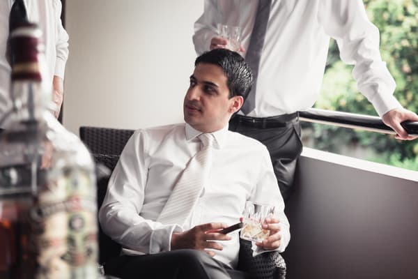 The groom, Pasha, dressed in a white shirt and tie, sits on a wicker chair holding a glass of whiskey and a cigar, while another man in a white shirt and tie stands nearby on a balcony with greenery visible outside. Engagement ceremony by She Said Yes.