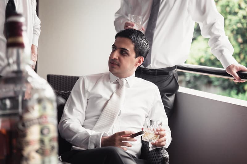 The groom, Pasha, dressed in a white shirt and tie, sits on a wicker chair holding a glass of whiskey and a cigar, while another man in a white shirt and tie stands nearby on a balcony with greenery visible outside.