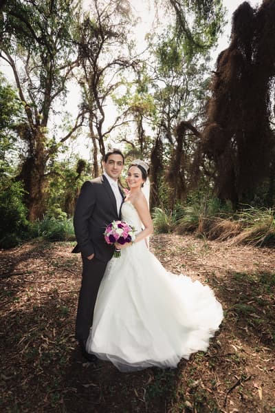 Pasha and Maryam pose together outdoors in a wooded area for couple portraits, with Maryam holding a bouquet of purple and white flowers. Wedding photographer and videographer by She Said Yes.