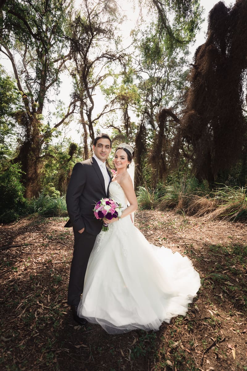 Pasha and Maryam pose together outdoors in a wooded area for couple portraits, with Maryam holding a bouquet of purple and white flowers.