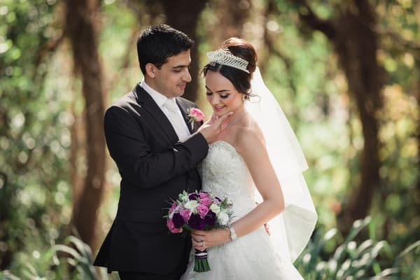 Pasha and Maryam pose together outdoors with Pasha gently touching Maryam's chin as she holds a bouquet of purple and white flowers. Destination wedding videographer by She Said Yes.