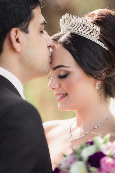 Pasha kisses Maryam on the forehead as she smiles with her eyes closed, wearing a tiara and diamond jewelry, holding a bouquet. Wedding photography coverage options by She Said Yes.