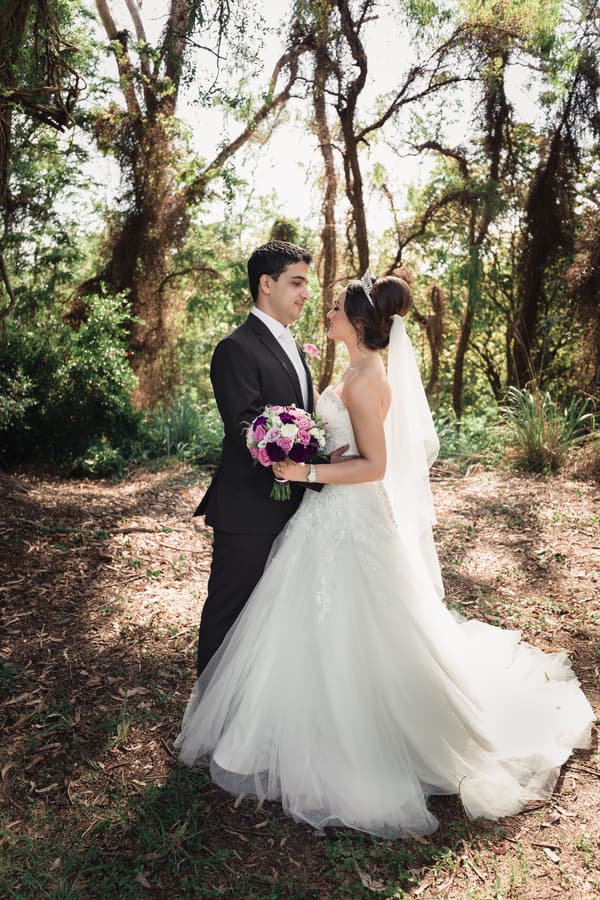 Pasha and Maryam stand facing each other outdoors surrounded by trees, with Maryam holding a bouquet of purple and white flowers.