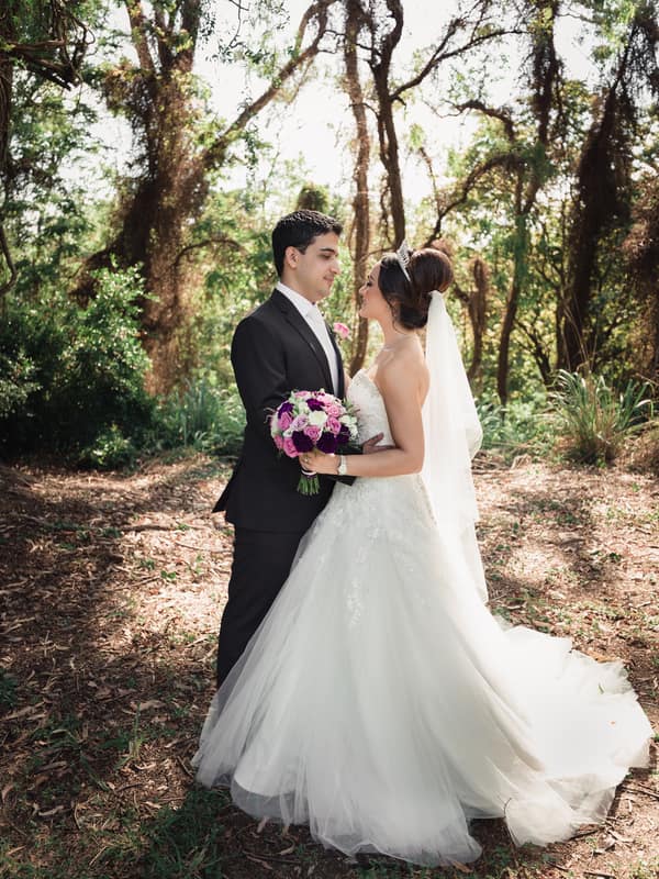 Pasha and Maryam stand facing each other outdoors surrounded by trees, with Maryam holding a bouquet of purple and white flowers.
