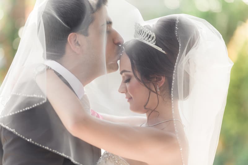 The groom kisses the bride's forehead as they embrace under her veil, the bride wearing a tiara and necklace.