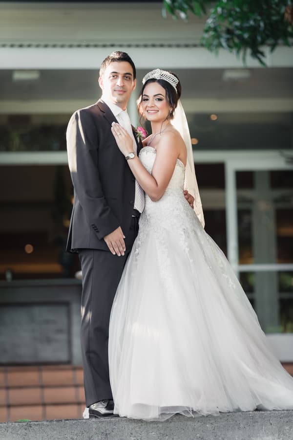 The bride Maryam in a white wedding gown and tiara stands next to the groom Pasha in a dark suit and light tie, posing together outside Hillstone St Lucia — The Quartyard.