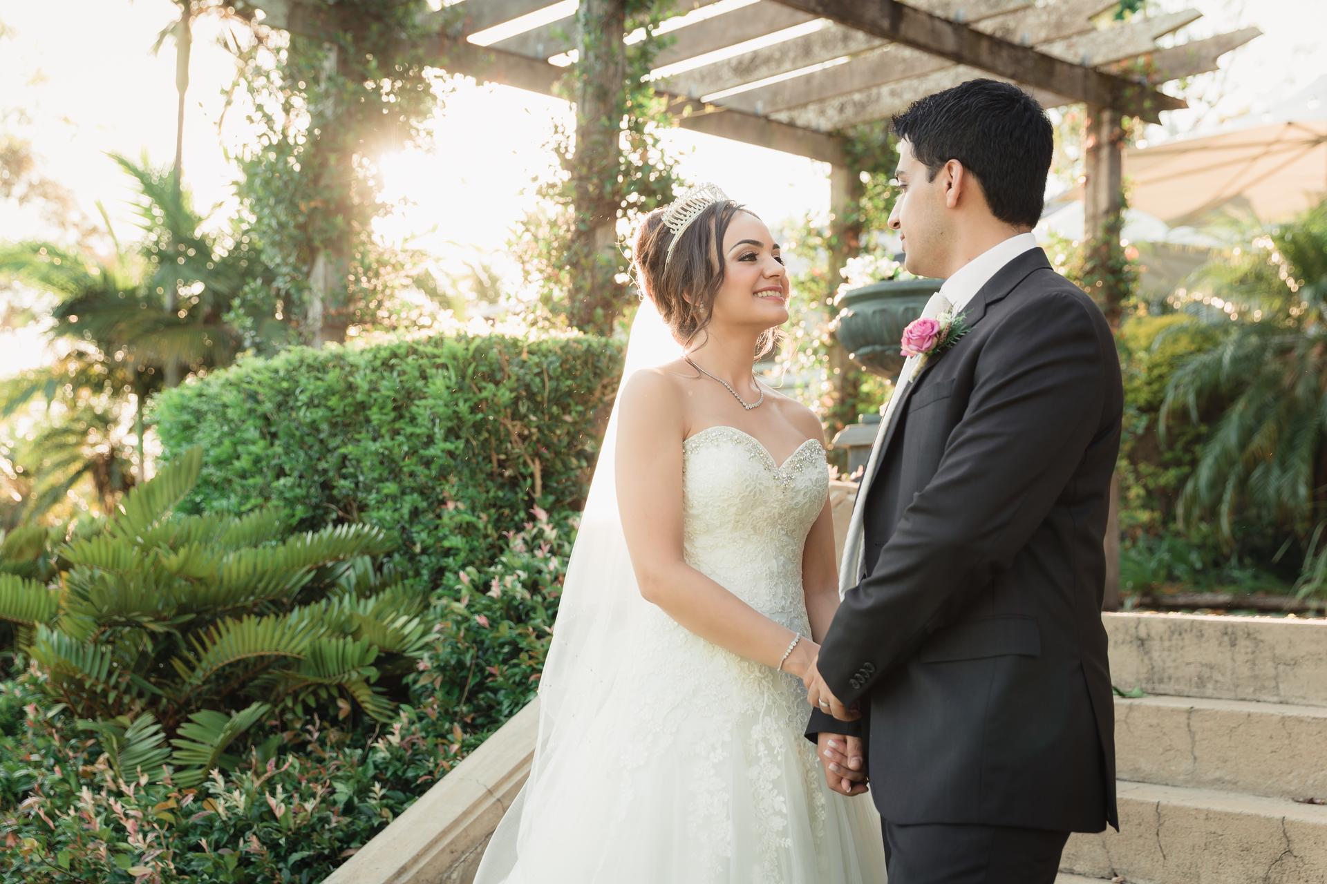 Bride Maryam and groom Pasha hold hands and look at each other outdoors at Hillstone St Lucia under a pergola with greenery in the background.