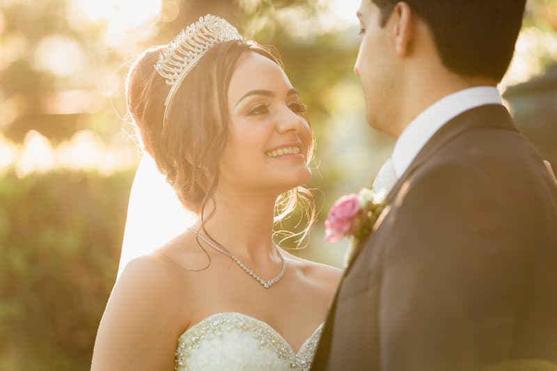 The bride Maryam wearing a tiara and necklace smiles at the groom Pasha, who is dressed in a suit with a pink boutonniere, in an outdoor setting with sunlight filtering through trees.