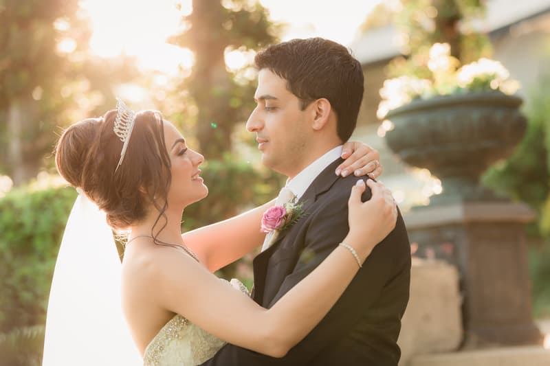 The bride and groom embrace outdoors with the bride wearing a tiara and veil and the groom in a black suit with a pink boutonniere.