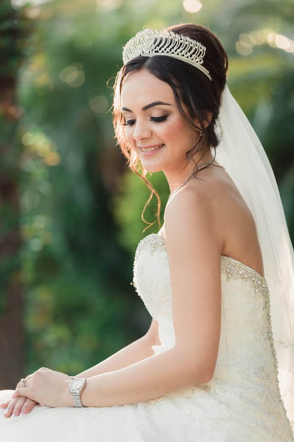 The bride Maryam in a strapless white wedding gown with a jeweled tiara and veil, seated outdoors with greenery in the background.