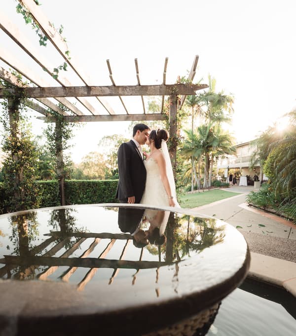 The bride Maryam and groom Pasha stand close under a wooden pergola at Hillstone St Lucia — The Quartyard, with their reflection visible in a nearby water feature.