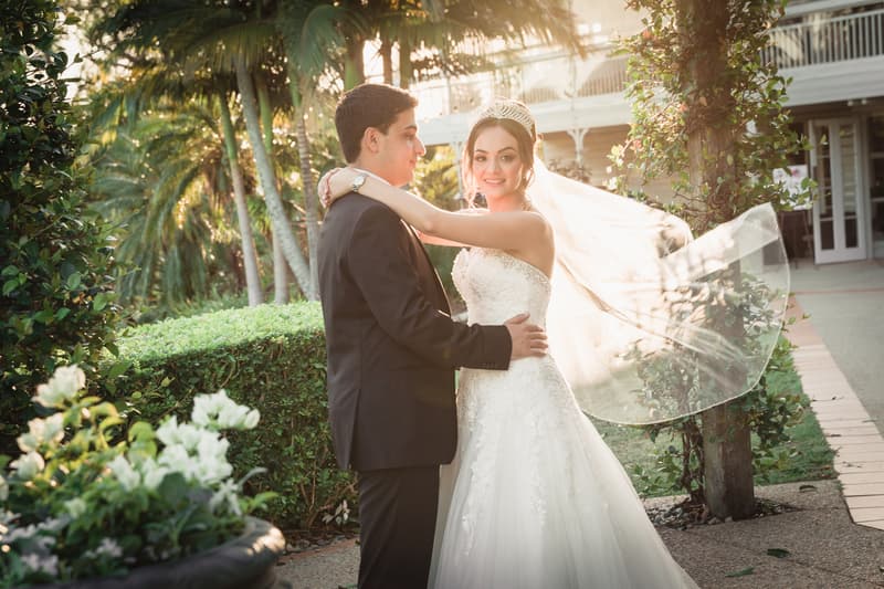The bride Maryam and groom Pasha pose together outdoors at Hillstone St Lucia in a garden area with greenery and palm trees, with Maryam's veil flowing in the breeze.