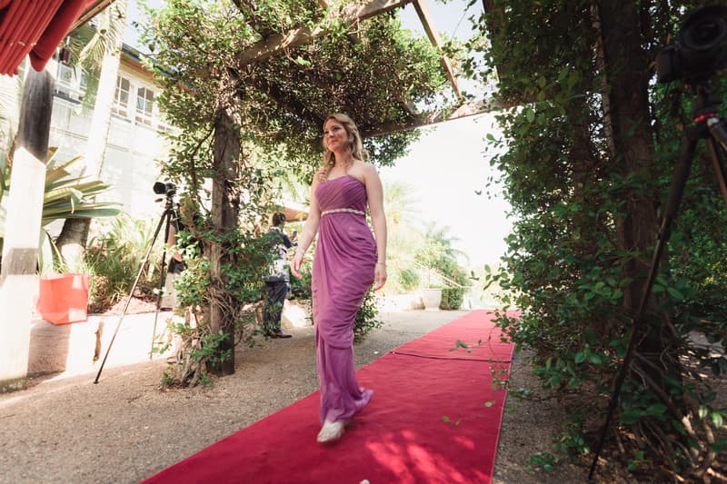 A bridesmaid in a purple dress walks down a red carpeted path under a wooden pergola covered with greenery at Hillstone St Lucia — The Quartyard.