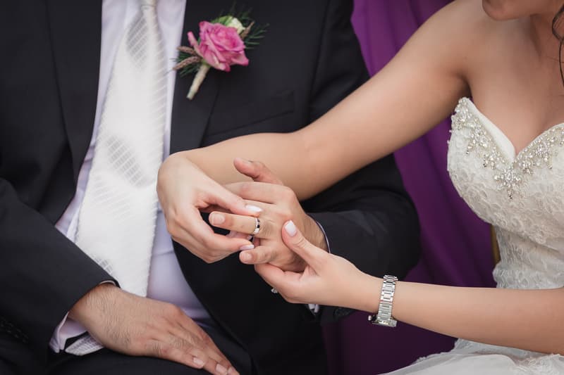 The bride places a wedding ring on the groom's finger; the groom wears a black suit with a white tie and a pink boutonniere, and the bride wears a white beaded wedding dress and a silver watch.