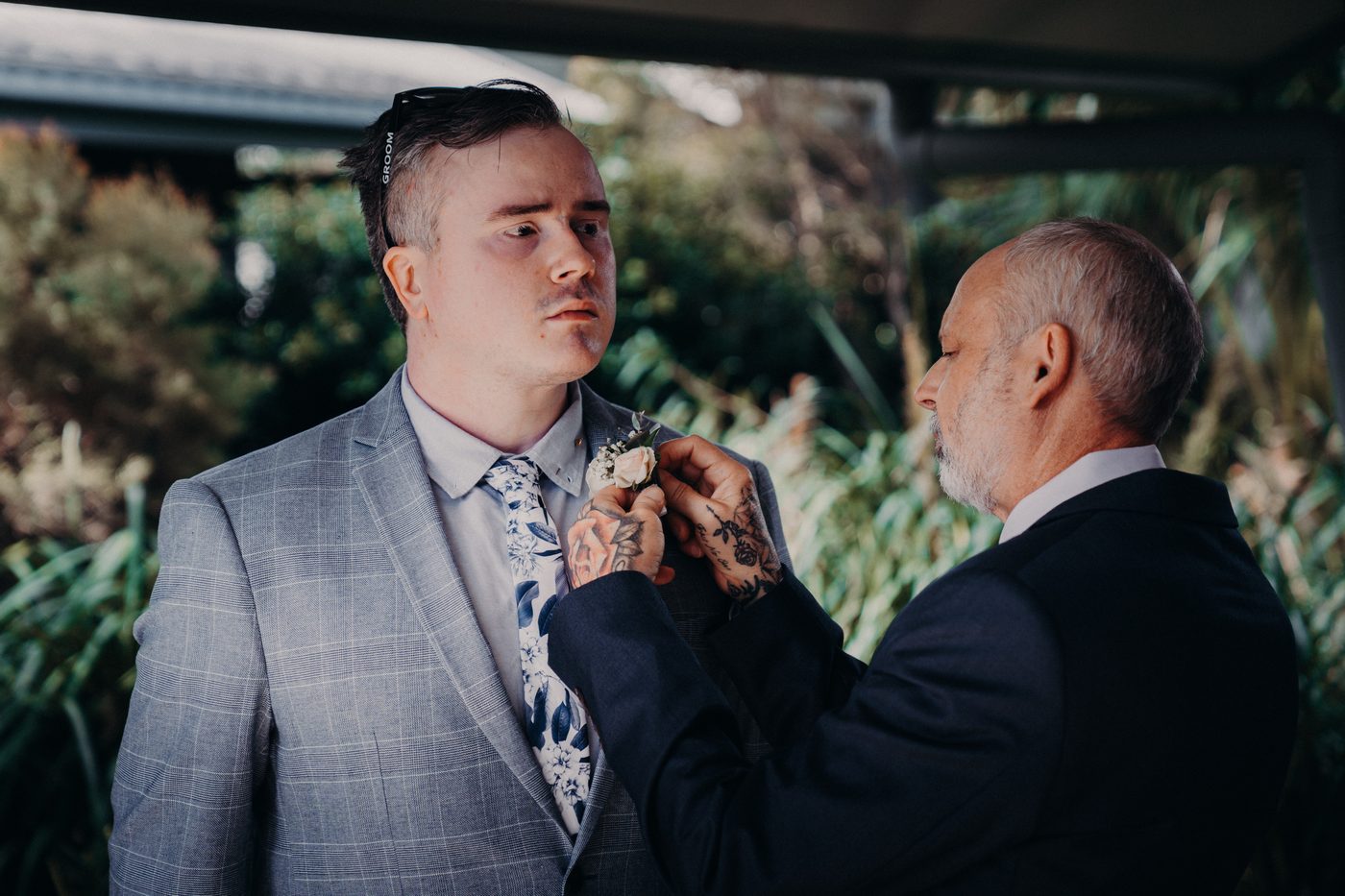 The groom has his boutonniere pinned by an older man, likely his father, under a covered outdoor area with greenery in the background. Sandstone point hotel wedding by She Said Yes.