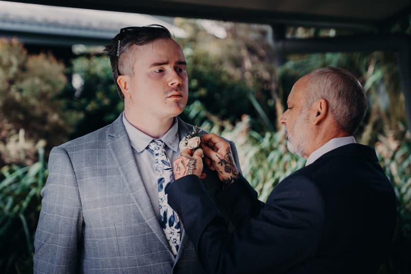 The groom has his boutonniere pinned by an older man, likely his father, under a covered outdoor area with greenery in the background at Sandstone Point Hotel.