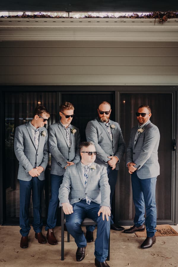 Dale and his groomsmen pose together wearing matching grey checked jackets, blue pants, floral shirts, and sunglasses at Sandstone Point Hotel.