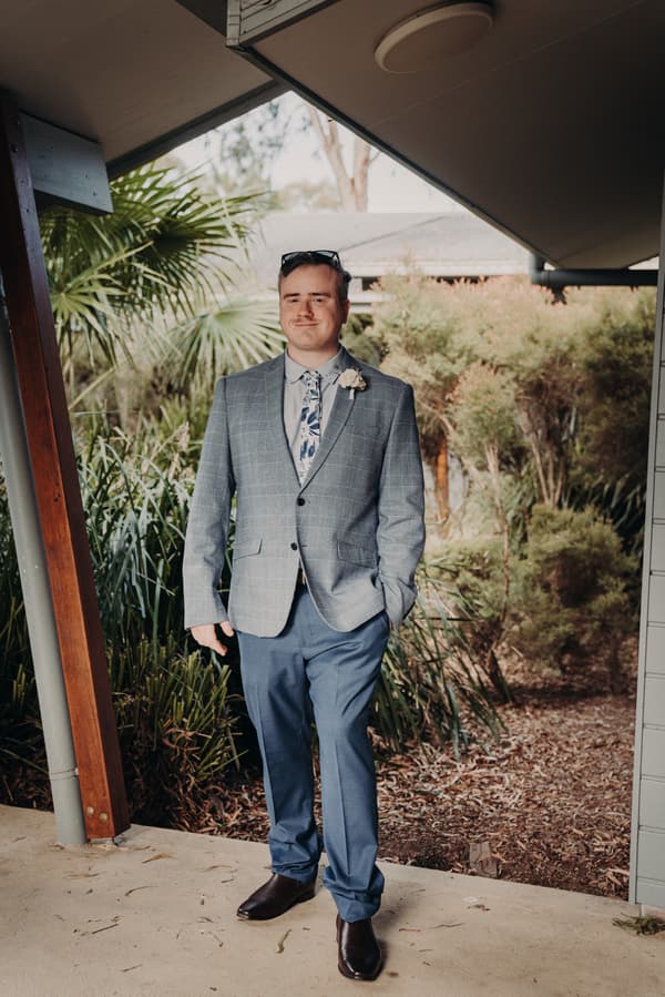 The groom stands alone under a covered outdoor area at Sandstone Point Hotel, wearing a grey checked blazer, blue trousers, floral tie, and a boutonniere.