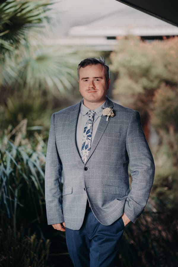 Dale, the groom, stands outdoors at Sandstone Point Hotel wearing a grey checked suit jacket, floral tie, and boutonniere.