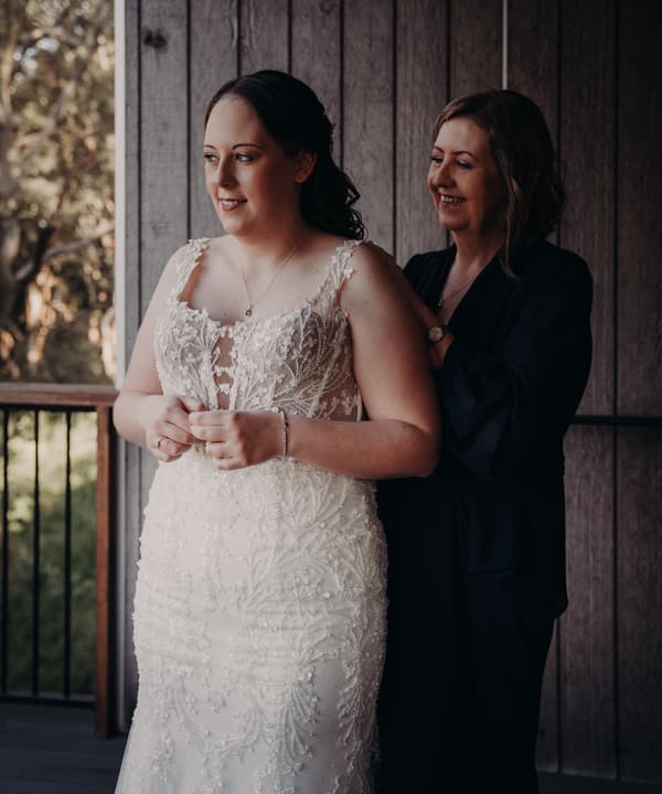 The bride Rebecca stands in a white lace wedding dress while the mother of the bride helps adjust the back of the dress at Sandstone Point Hotel — Rustic Arbour.