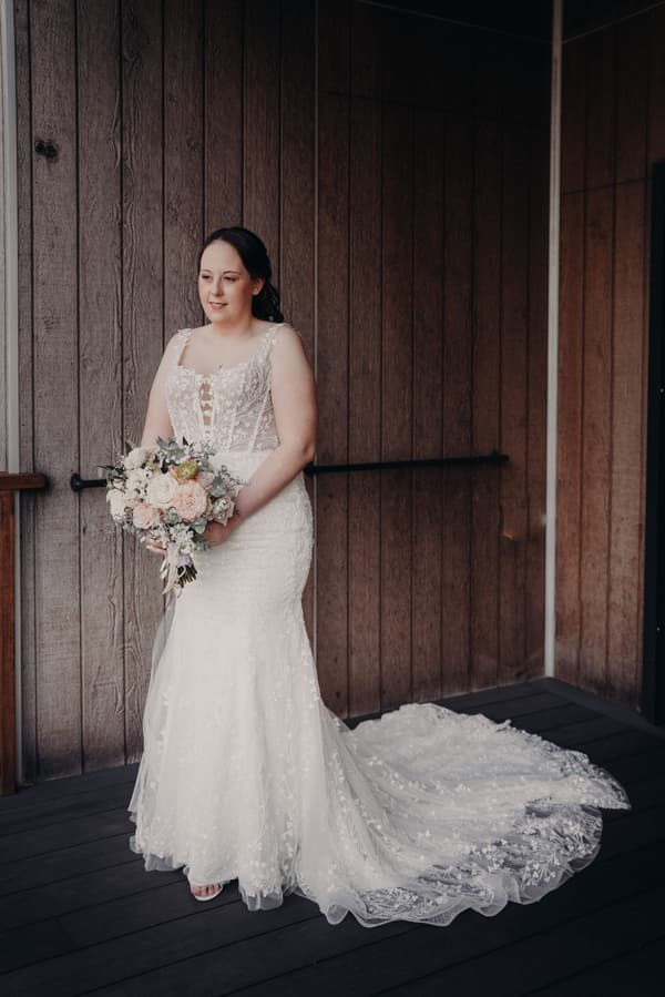 The bride Rebecca stands alone holding a bouquet of flowers against a wooden wall at Sandstone Point Hotel.