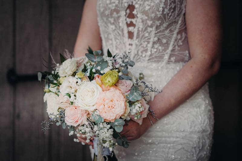 The bride holds a bouquet of mixed flowers including white and pale pink roses, greenery, and other floral accents, wearing a detailed lace wedding dress. Candid wedding photographer by She Said Yes.