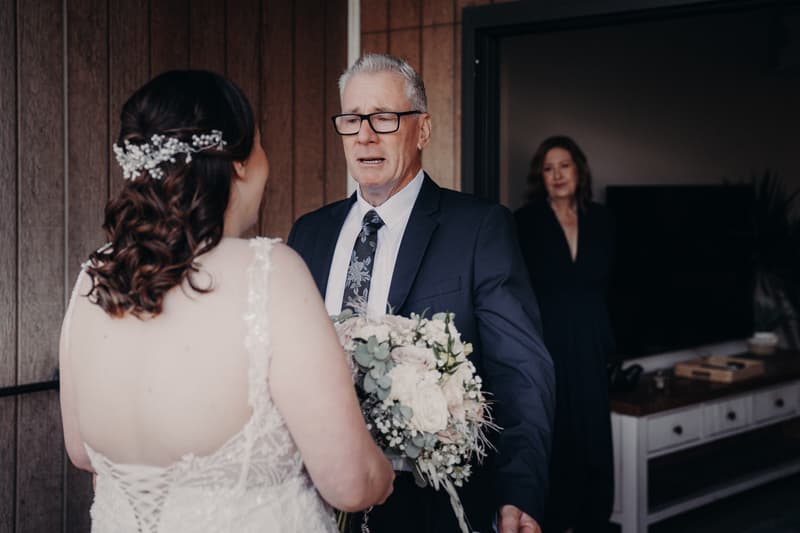 The bride holds a bouquet facing an older man in a suit and tie, who appears emotional, while an older woman stands in the background inside a room at Sandstone Point Hotel.