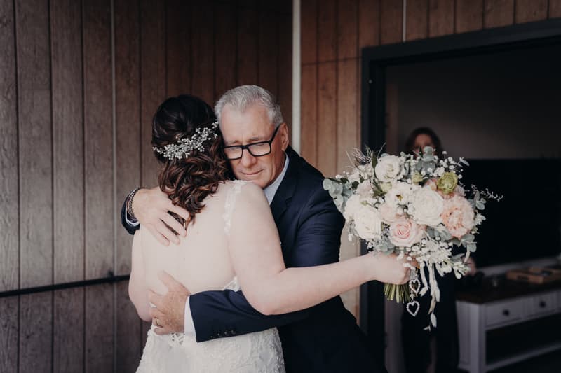 The bride Rebecca embraces an older man, likely her father, who is wearing glasses and a dark suit, inside a room at Sandstone Point Hotel.