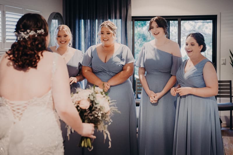 The bride Rebecca, holding a bouquet, faces four bridesmaids dressed in matching blue gowns inside a room with a window and curtains.