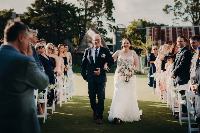 The bride Rebecca walks down the aisle arm-in-arm with an older man, likely her father, at Sandstone Point Hotel — Rustic Arbour, while guests stand on either side watching.