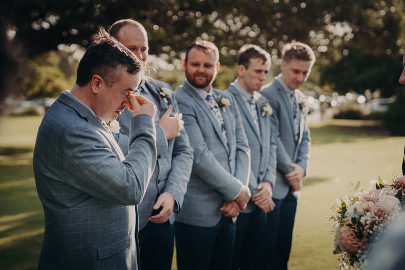 The groom and his groomsmen stand in a line outdoors at Sandstone Point Hotel — Rustic Arbour, with the groom wiping his eye and the others looking forward.