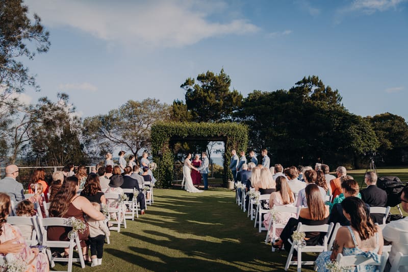 Bride Rebecca and groom Dale stand under a rustic arbour at Sandstone Point Hotel exchanging vows during their outdoor wedding ceremony, with bridesmaids and groomsmen standing on either side and guests seated in rows watching.