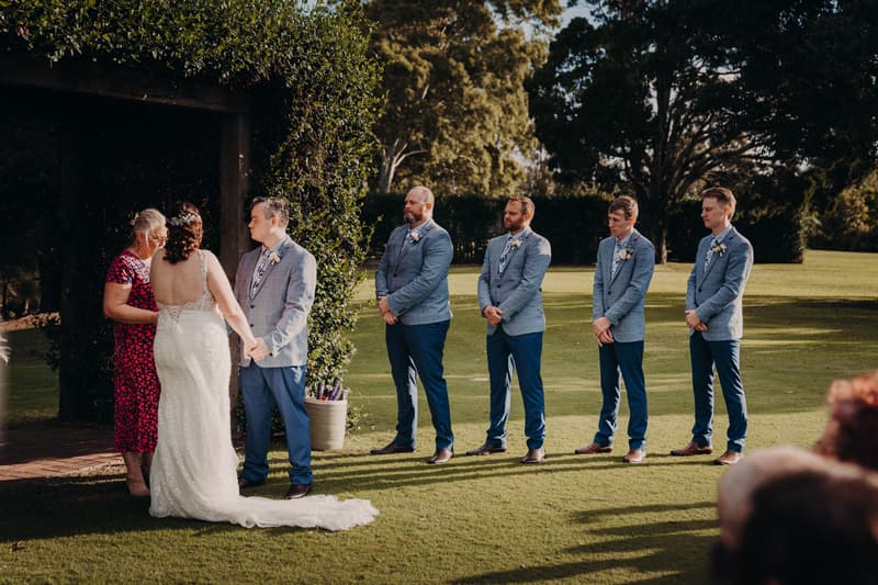 Rebecca and Dale hold hands facing the officiant during their wedding ceremony at Sandstone Point Hotel — Rustic Arbour, with four groomsmen standing in a line to the right on the grass.