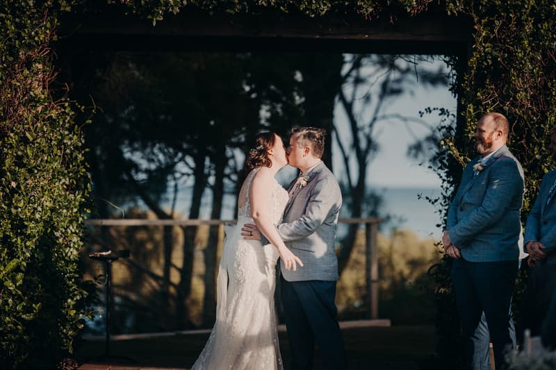 Rebecca and Dale kiss at the ceremony stage under a rustic arbour at Sandstone Point Hotel, with a groomsman standing to the side.